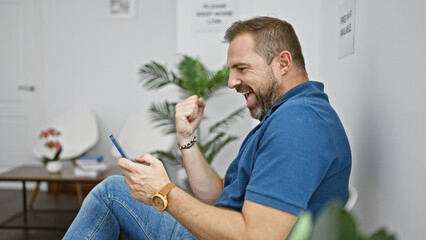 Handsome middle-aged hispanic man with grey hair laughing indoors, holding a smartphone in a bright room.