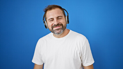 Smiling bearded man in white shirt with headphones against a blue wall outdoors.