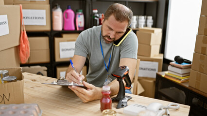 Mature hispanic man with grey hair managing donations in a warehouse, writing on a clipboard.