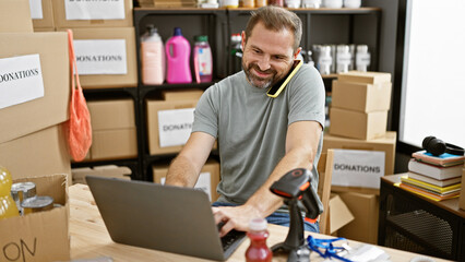 Handsome middle-aged man multitasking on a laptop with a phone at a busy donation center warehouse.