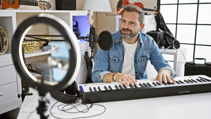 Handsome middle-aged hispanic man playing keyboard in well-equipped home music studio with recording equipment.