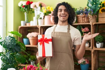 Hispanic man with curly hair working at florist shop holding gift celebrating achievement with happy smile and winner expression with raised hand