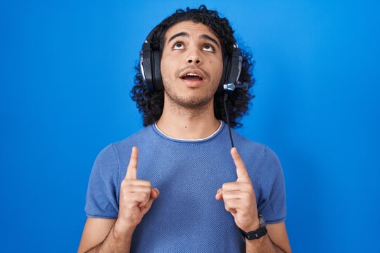 Hispanic man with curly hair listening to music using headphones amazed and surprised looking up and pointing with fingers and raised arms.