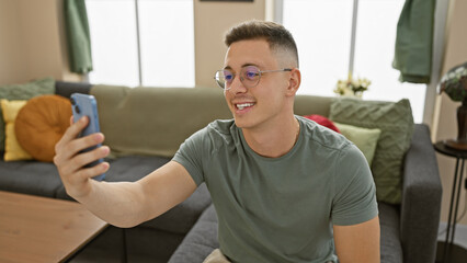 Handsome young man taking a selfie indoors with his phone, casually sitting in a modern living room.