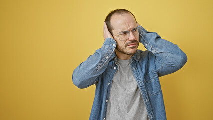 A bald man with a beard wearing glasses and a denim shirt covers his ears against a yellow background.