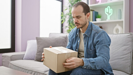 Hispanic bald man with beard holding box sitting on grey sofa in a modern living room