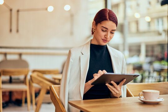 An elegantly dressed woman, using a digital tablet while sitting at the cafe, drinking a cup of coffee.
