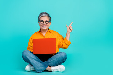 Full body photo of old grandmother sit floor with laptop point finger empty space courses app isolated on aquamarine color background