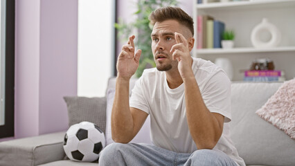 A hopeful young man with a beard sitting indoors, crossing his fingers in a living room with a...
