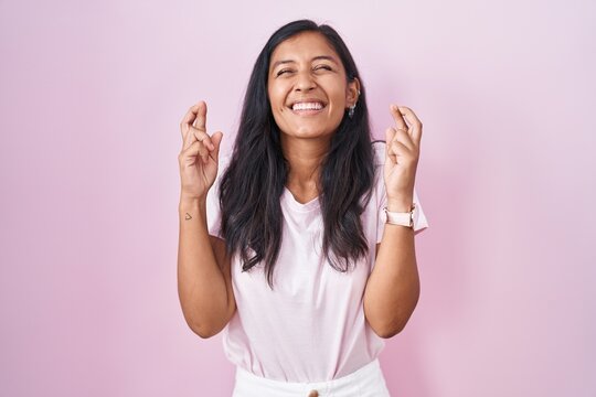 Young hispanic woman standing over pink background gesturing finger crossed smiling with hope and eyes closed. luck and superstitious concept.