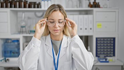 A young blonde woman in a lab coat adjusts her glasses indoors at a laboratory