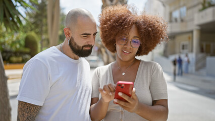 Beautiful couple, laughing and confidently standing together on a city street, smiling and bonding over shared messages on their smartphone