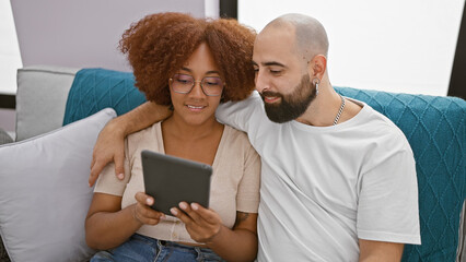 Beautiful smiling couple sitting together at home, confidently enjoying online bonding over touchpad, radiating joy in their lovely indoor living room relationship