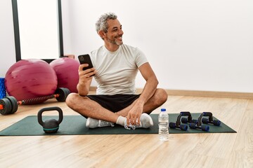 Middle age grey-haired man sitting on yoga mat using smartphone at sport center