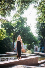 A beautiful slender aged woman with long blond hair, in a black long sexy dress, walks barefoot along a picturesque fountain in a city park, on a bright sunny day.