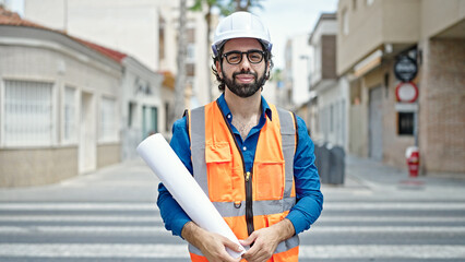 Young hispanic man architect smiling confident holding blueprints at construction place - obrazy, fototapety, plakaty