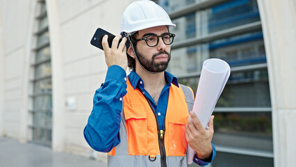 Young hispanic man architect holding blueprints listening to voice message by smartphone at...