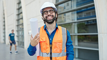 Young hispanic man architect smiling confident holding blueprints at construction place