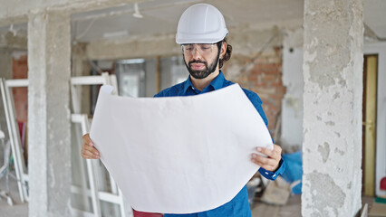 Young hispanic man architect reading house plans standing at construction site