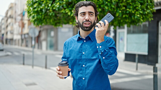 Young hispanic man sending voice message by smartphone drinking coffee at street