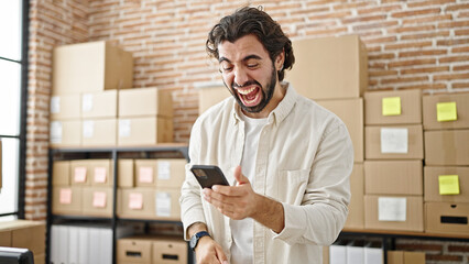 Young hispanic man ecommerce business worker using smartphone celebrating at office