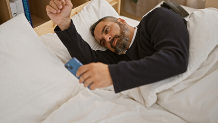 A mature hispanic man with grey beard lies in bed, casually using a smartphone in a cozy room.