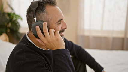 Mature hispanic man with beard and grey hair smiling while listening to headphones in a cozy home interior.