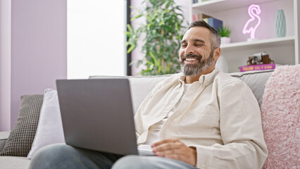 Mature hispanic man with a beard and grey hair smiling while using a laptop on a sofa.