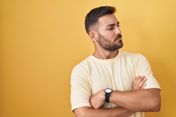 Handsome hispanic man standing over yellow background looking to the side with arms crossed...