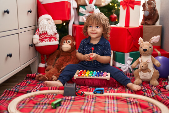 Adorable hispanic toddler playing xylophone sitting on floor by christmas gifts at home