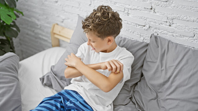 Adorable blond boy sitting in bedroom, scratching itchy arm rash - indoor allergy problem or eczema? child's health at stake in home environment!