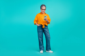 Full body length photo of gray haired middle age woman wear orange shirt jeans surfing social networks isolated on blue color background