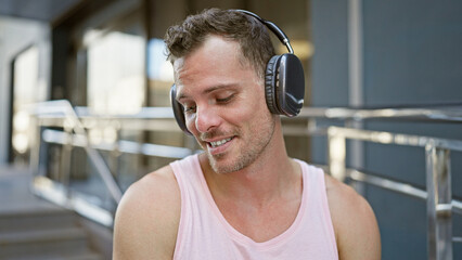 A smiling young hispanic man with headphones enjoys music in an urban street setting, exuding a relaxed vibe.