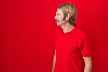 Caucasian man with mustache standing over red background looking to side, relax profile pose with...
