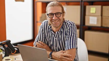 Smiling man with glasses in striped shirt seated at a desk with a laptop in a warehouse office.