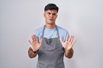 Hispanic young man wearing apron over white background moving away hands palms showing refusal and...