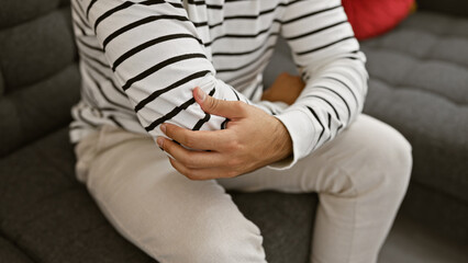 Hispanic man's hands gripping elbow in pain, sitting on a cozy sofa at home, resting and looking for comfort. adult male suffering from an indoor relaxed lifestyle, likely injury causing ache.