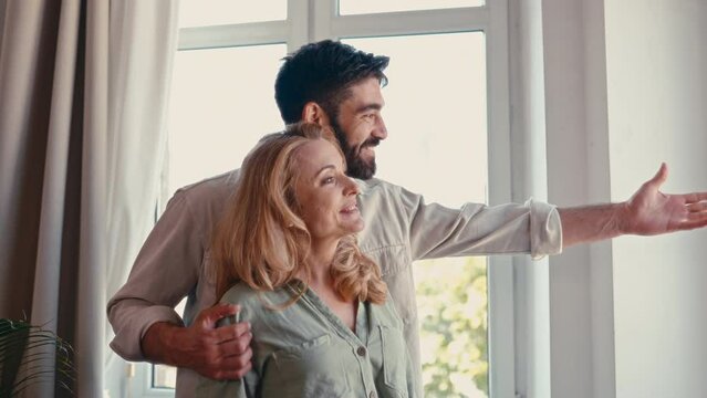 Beautiful adult couple hugging while standing by the window in the living room at home, pointing with his hand at something on the street outside the window, spending time together romantically.
