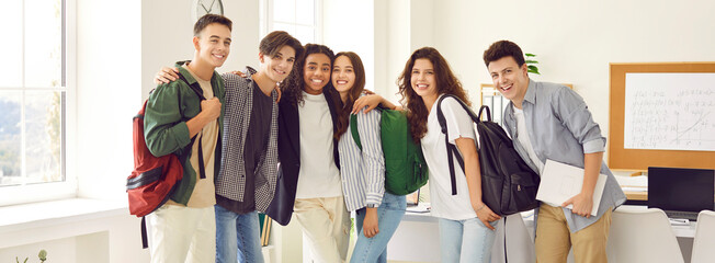 Portrait of a happy cheerful smiling high school students group standing in classroom together and hugging. Friendly guys and girls standing in a row indoors. Education and back to school concept.