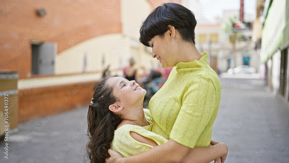 Wall mural Confident mother and daughter share a joyous, smiling hug on a sunny street, strengthening their lovely relationship outdoors