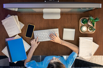 A schoolgirl with a smartphone sits in front of a computer, studying online.