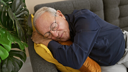 Cozy indoor snapshot, exhausted elder man with grey hair in glasses resting in comfort, lounging on the sofa at home, surrendering to a well-deserved sleep after a tiring day
