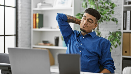 Young latin man business worker using touchpad and laptop stretching back at the office
