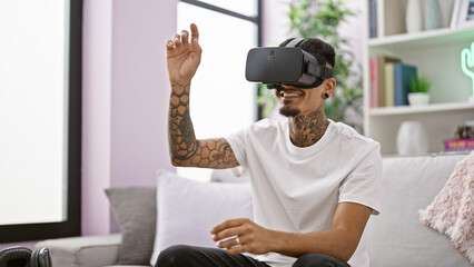 Cheerful tattooed young latin man, all smiles, chilling on his comfy sofa at home, confident. totally immersed in a new level of gaming, using virtual reality glasses, tech at its best. - obrazy, fototapety, plakaty