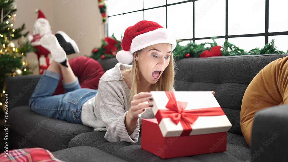 Sticker Young blonde woman unpacking gift lying on the sofa by christmas tree at home