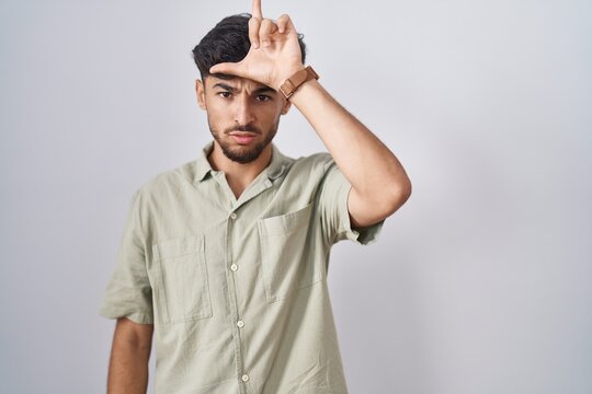 Arab man with beard standing over white background making fun of people with fingers on forehead doing loser gesture mocking and insulting.