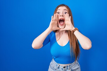 Redhead woman standing over blue background smiling cheerful playing peek a boo with hands showing...