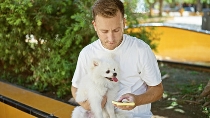 Young caucasian man with dog sitting on bench using smartphone at park
