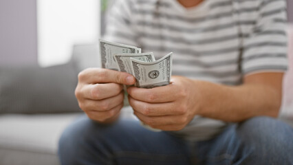 Handsome young caucasian man counting dollars on sofa at home, managing personal finance in the...
