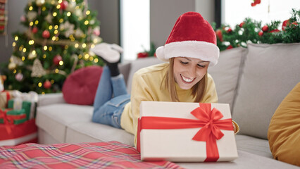 Obraz na płótnie Canvas Young blonde woman unpacking gift lying on sofa by christmas tree at home
